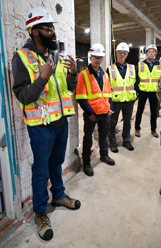 A black man in an orange safety vest and white hard hat stands in a line with three white men also in vests and hardhats.