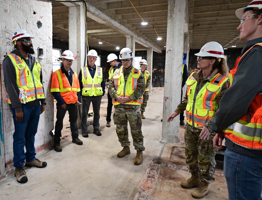 A group of one black man, one white woman and six white men, all in orange safety vests and white hard hats, stand facing each other talking.