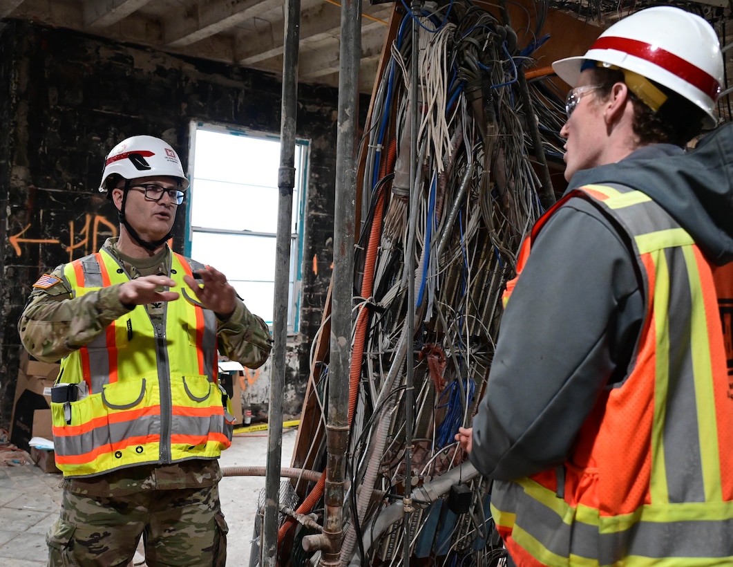 Two white males in orange construction vests and white hard hats stand facing each other as they talk.