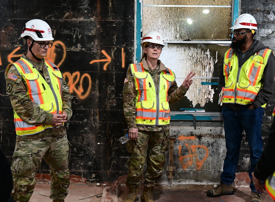 A white man in Army camo with a white hard hat stands next to a white female in Army camo with a white hard hat, who stands next to a black man in blue jeans, dark grey jacket and orange safety vest in a room under construction.