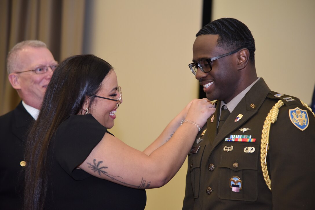 Photo is of a woman pinning US Army captain rank to the lapel of her husbands Army uniform.