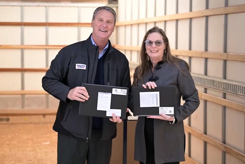 A man and a woman stand inside a trailer smiling and holding laptops to be donated.