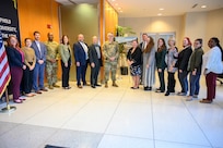 A row of civilian and military stand in front of flags in a lobby