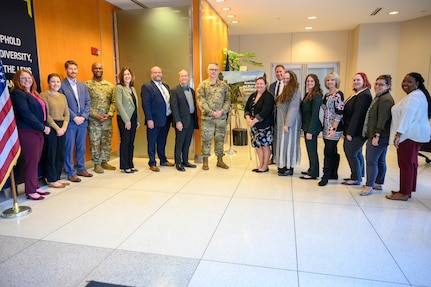 A row of civilian and military stand in front of flags in a lobby