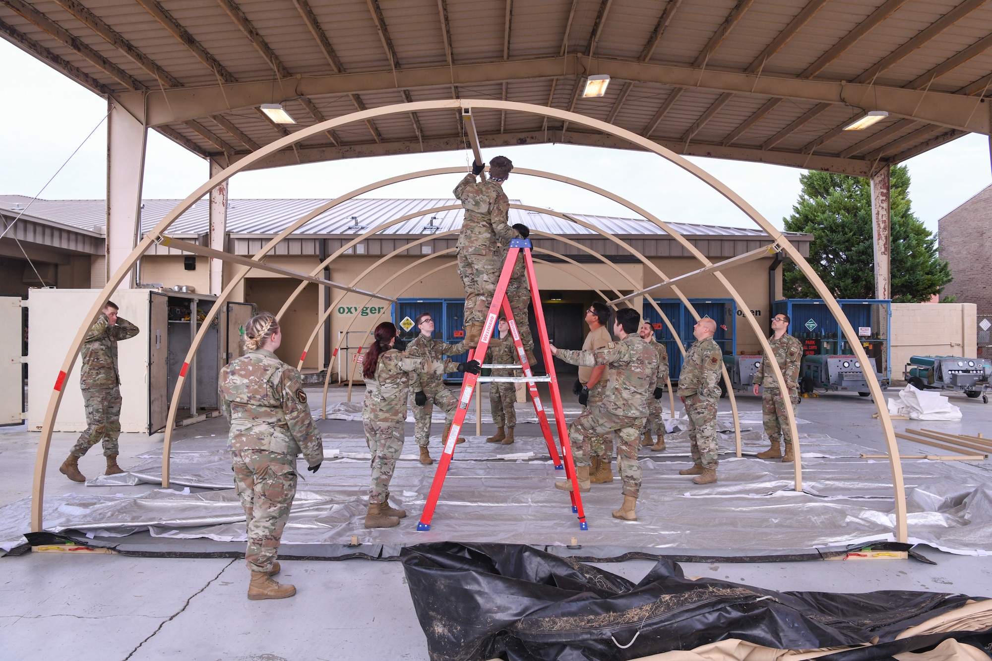 People assemble a tent frame, one man stands on a ladder to help.