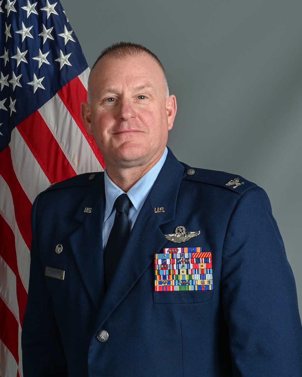 Middle age man in blue uniform stands in front of American flag with grey studio background