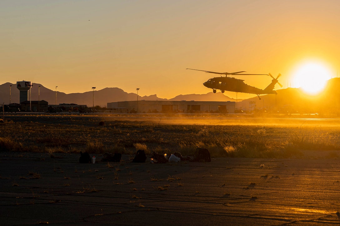 A helicopter prepares to land on a tarmac as the sun beams from right with silhouettes of mountains to the left.