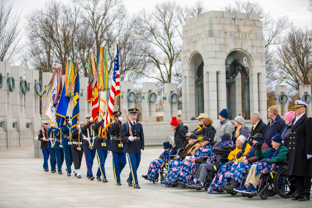 Soldiers in ceremonial uniform carrying flags walk in formation in front of seated veterans and their loved ones at a memorial.