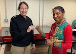 A woman in a Navy uniform with three silver stars on her shoulders hands a small coin to another woman in a red and green sweater. both are looking into the camera and smiling