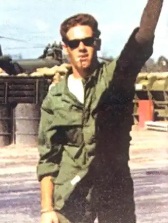 A man in a flight suit poses for a photo with one hand in the air.