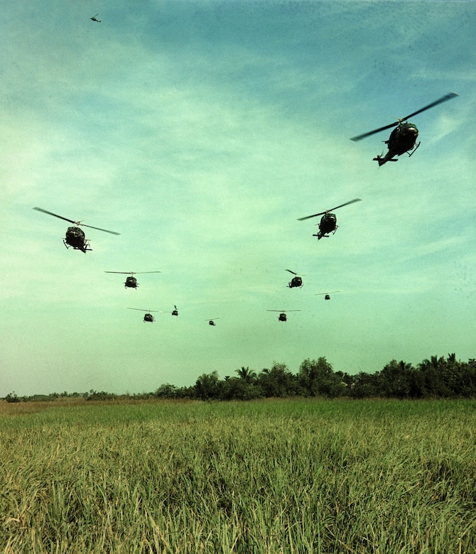 Ten helicopters fly above a grassy field.