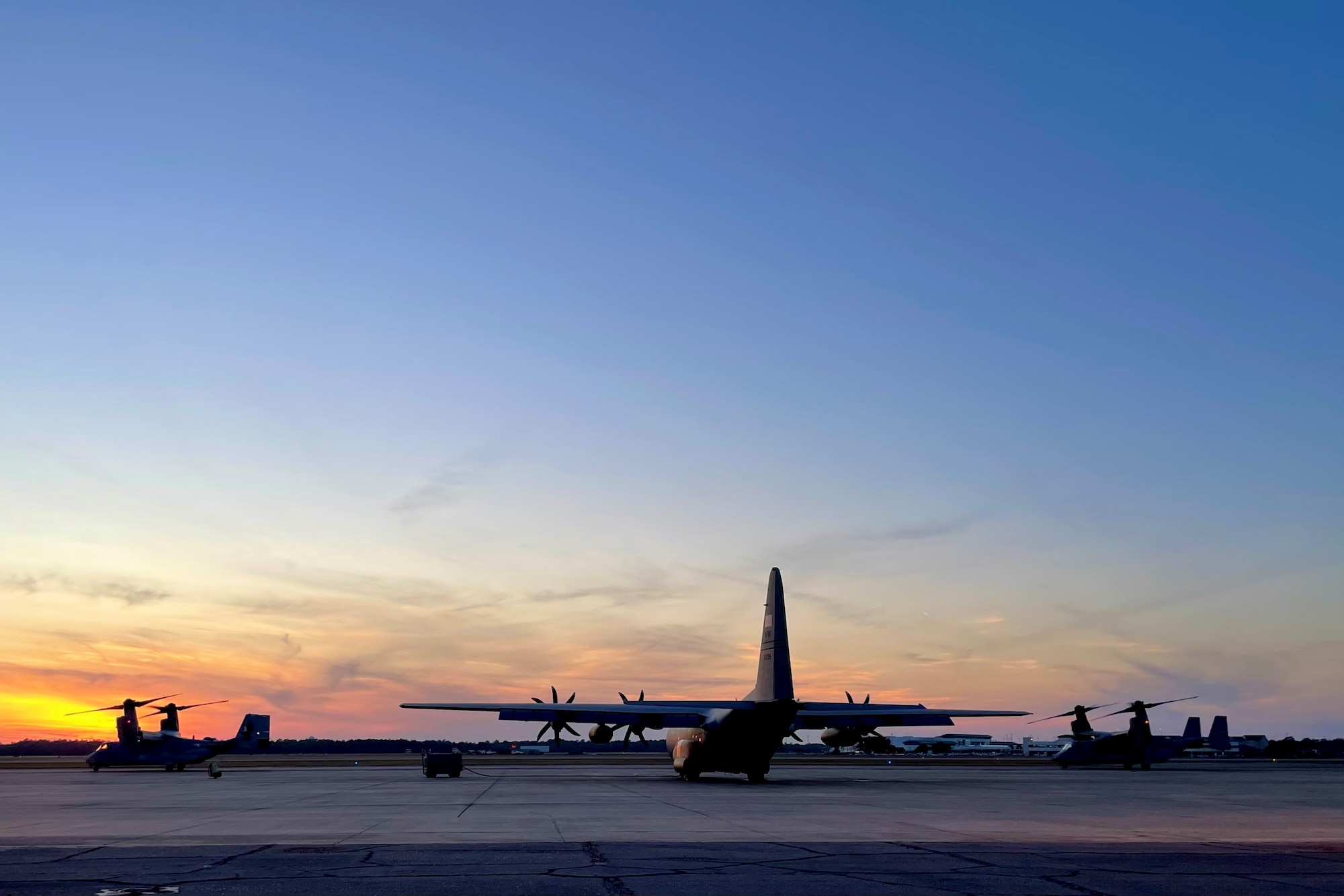 a C-130 is parked on a flight line during sunset while two MC-22s prepare to taxi