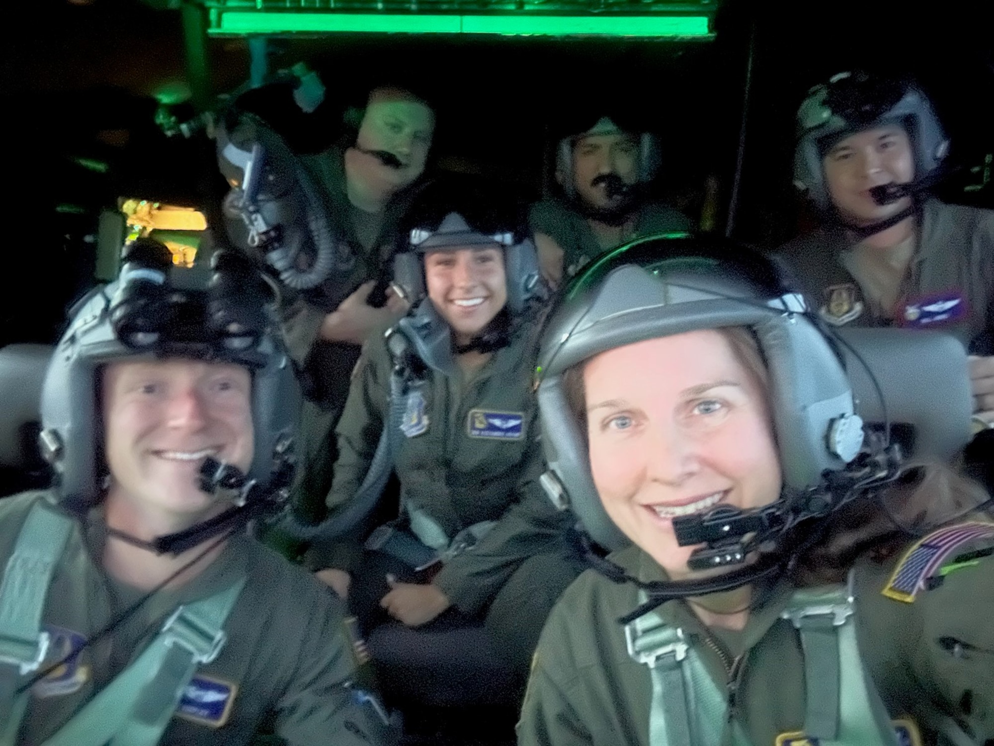 a group of people in green flight suits and helmets smile for a selfie inside an airplane cockpit