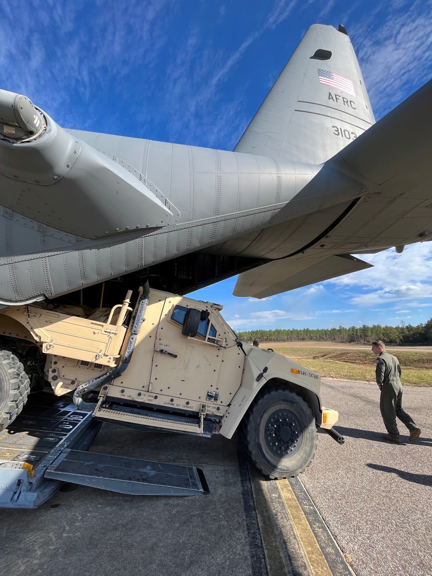 a man in a green flight suit walks behind the open ramp of an aircraft as a large vehicle is loaded inside
