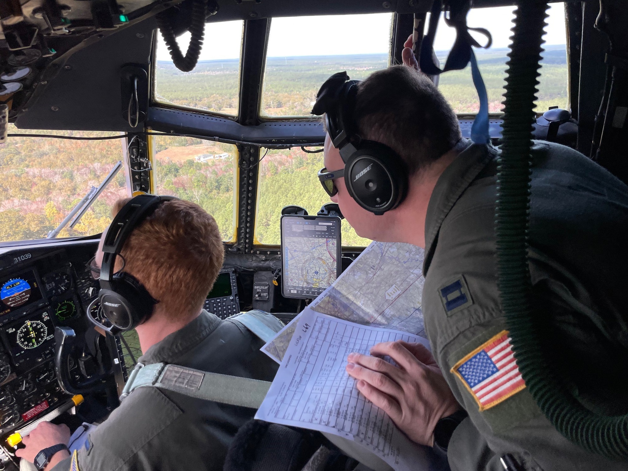 two men in green military uniforms look out over a cockpit during flight