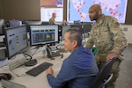 White man sits in front of a computer screen with a black man standing behind him