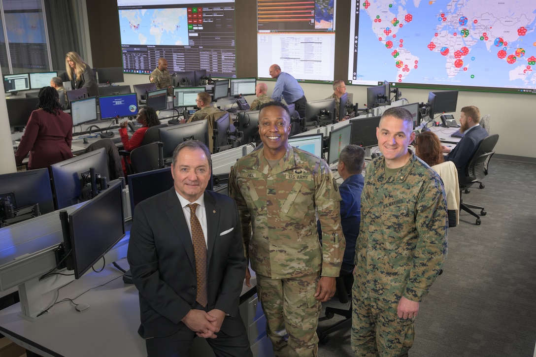 Two white men and one black man stand side by side in the front of a big room with a map projected on the back wall