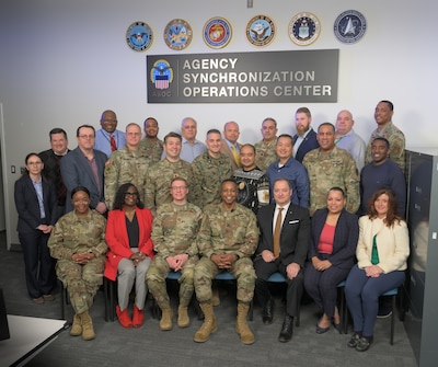 A large group of people pose for a picture under a sign that says Agency Synchronization Operations Center