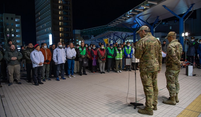 Two military men stand in front of a crowd at night.
