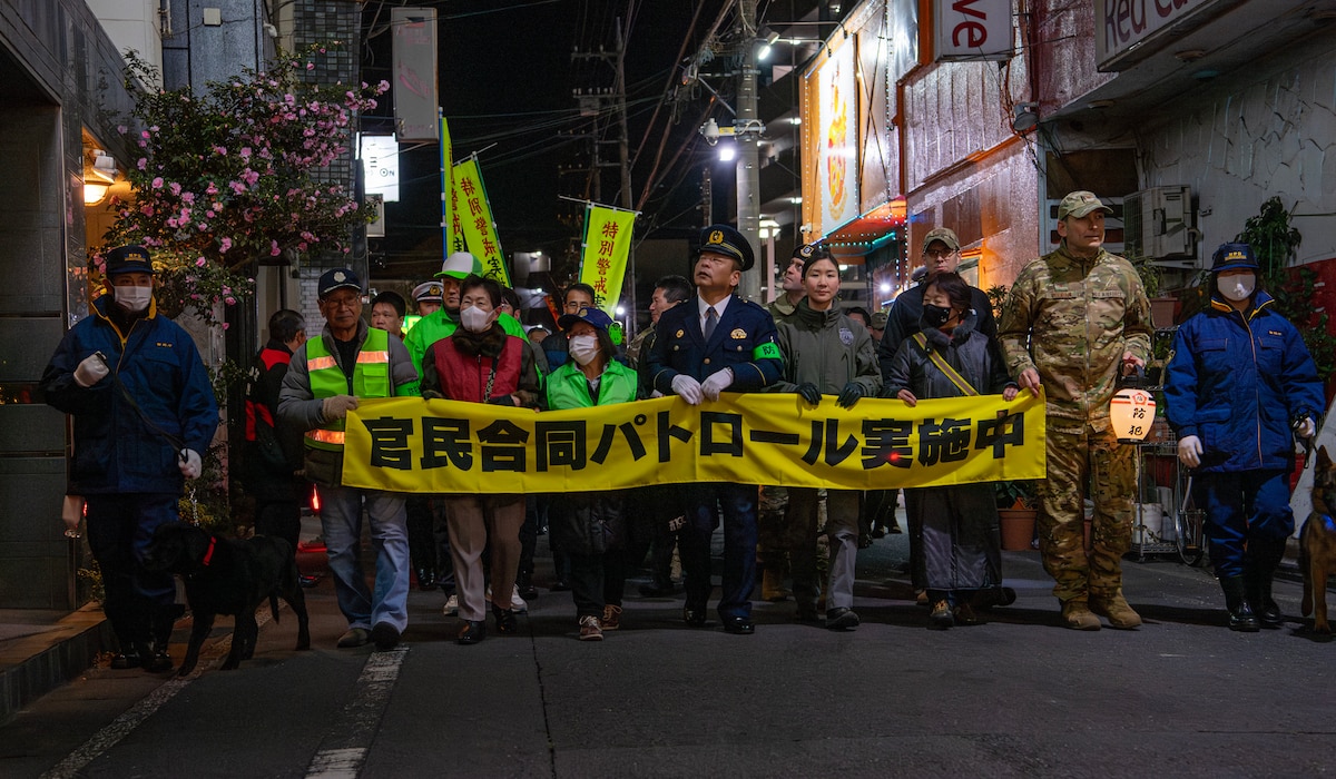 Military members, police, and local community members hold a banner while walking on a street.