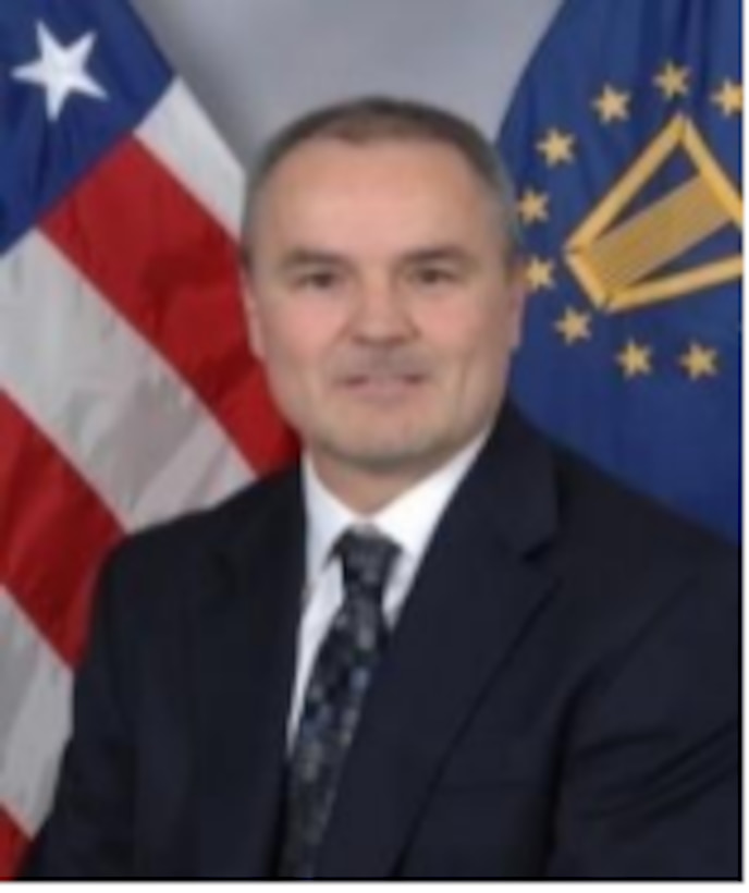 Headshot of person wearing a black suit in front of two flags.