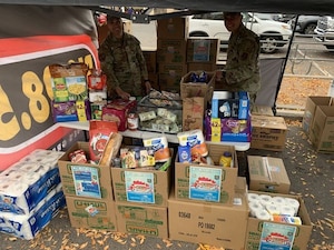 Two guardsmen stand under a tent with boxes full of supplies.