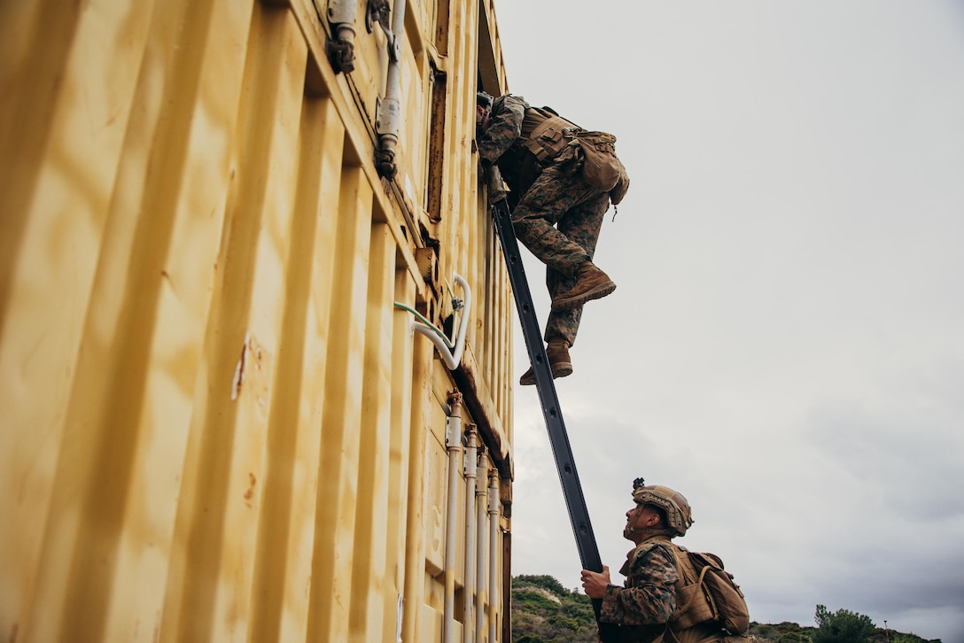 A Marine climbs a ladder to look into a metal building during training as a second Marine looks up from the bottom of the ladder.