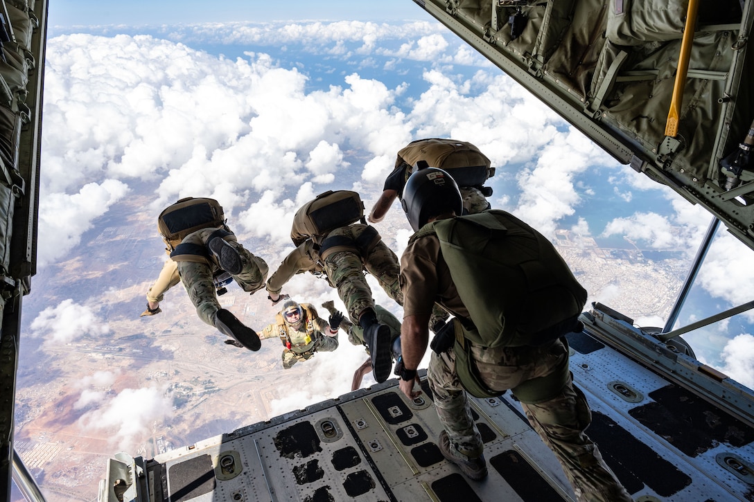 A group of airmen jump from a military aircraft during daylight. Clouds are visible from beneath the aircraft.