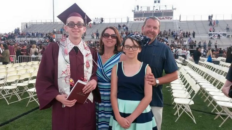 A man in a maroon cap and gown stands with a woman in a blue strip dress, a young girl in a blue and black strip dress, and a man in a blue shirt with white folding chairs in the background.