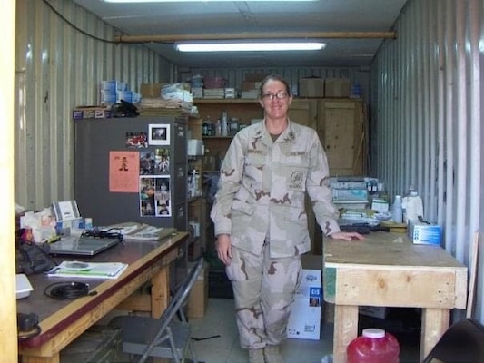 A woman in a military uniform stands with wooden tables on her left and right and a grey file cabinet in the background.