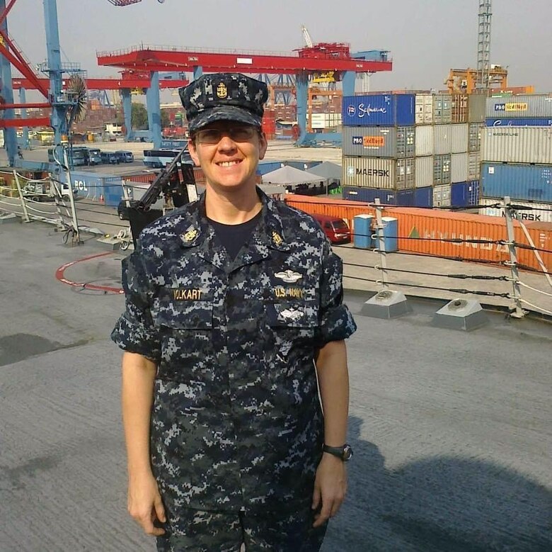 A woman in a blue Navy uniform stands in front of cargo on a ship.