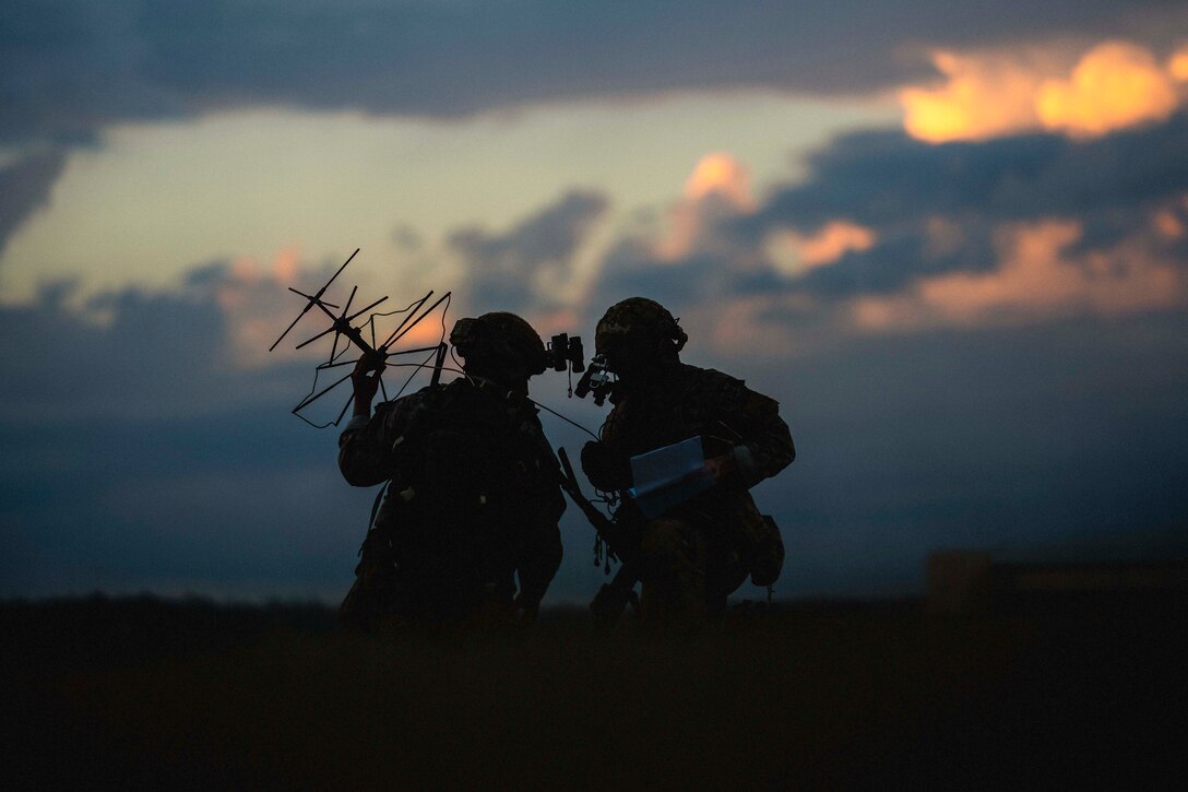 A soldier in tactical gear looks at device as another holds up an antenna as shown in silhouette.