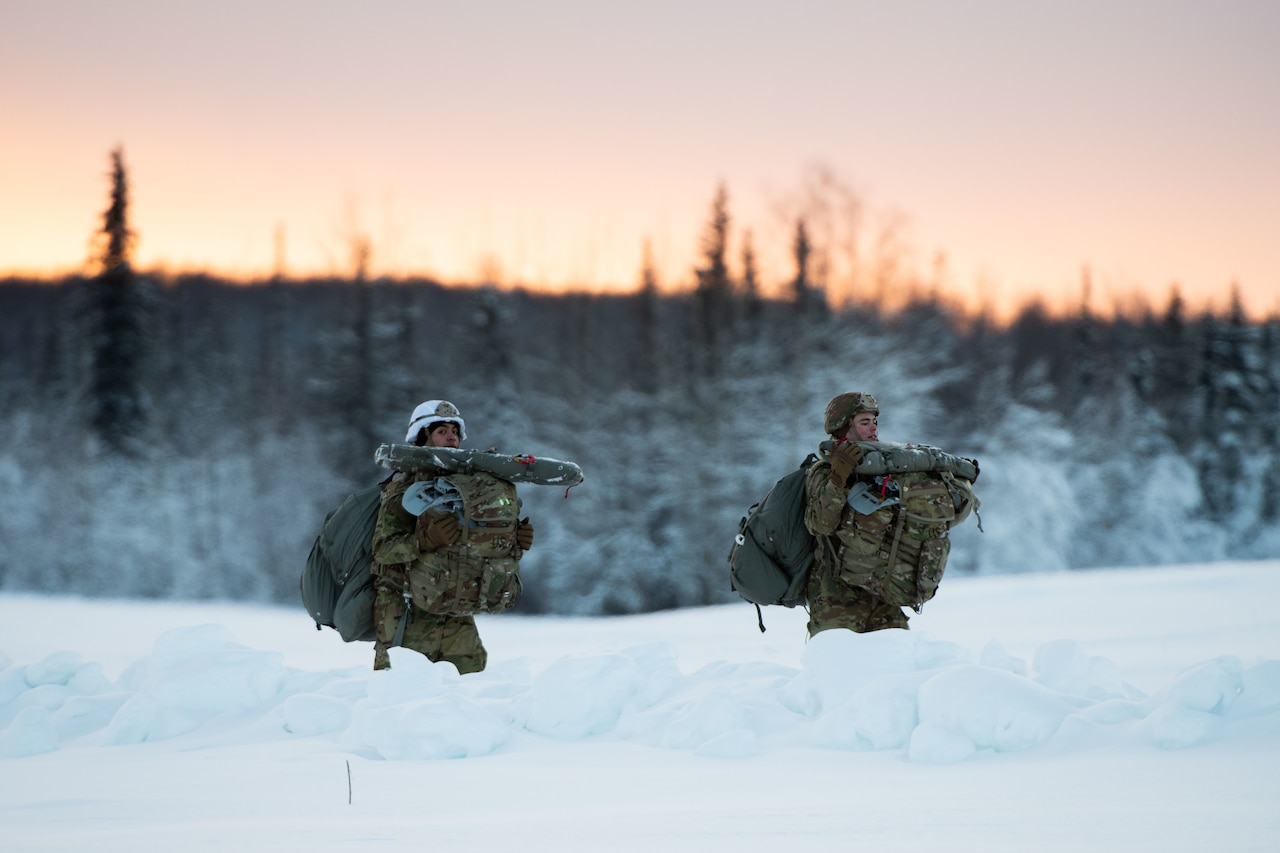 Two soldiers walk in the snow carrying gear.
