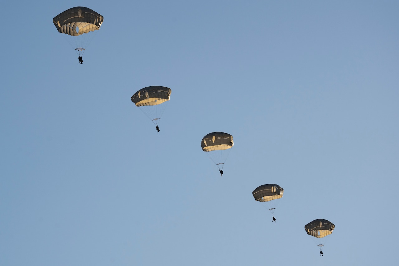 Soldiers descend with parachutes in a blue sky.
