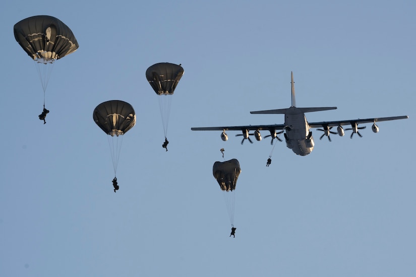 Soldiers descend with parachutes with an aircraft in the background.