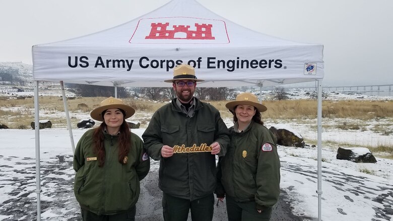 Three park rangers stand under a US Army Corps of Engineers tent, stand in front of the Dalles Lock & Dam with snow all around.