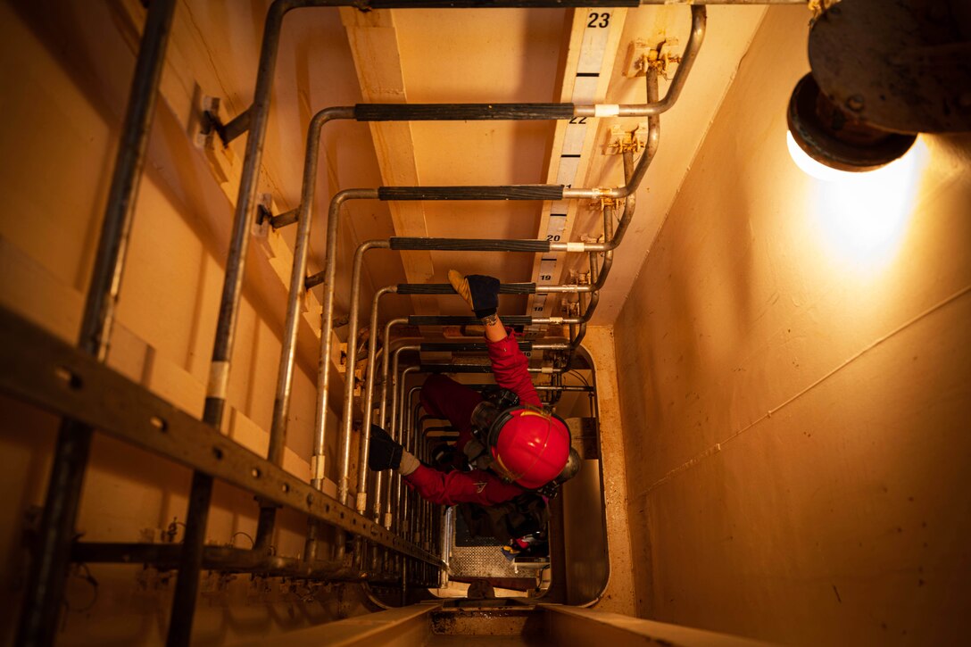 Seen from above, a wall lamp lights a sailor climbing halfway up a ladder in a narrow, enclosed space.