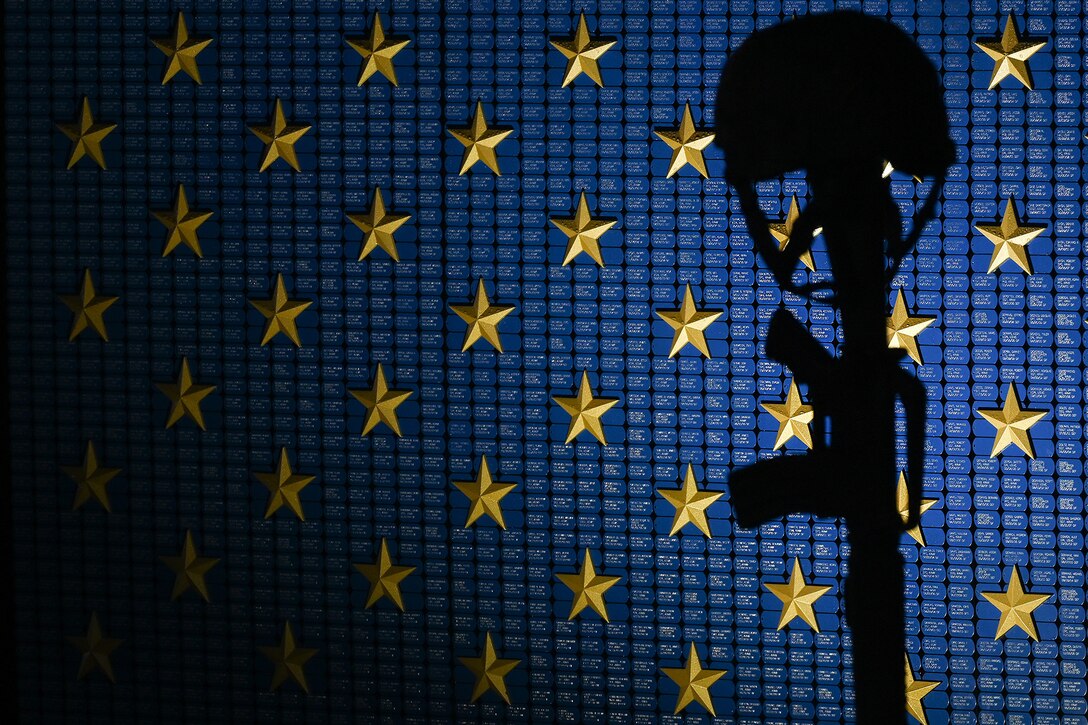 A battlefield cross is silhouetted against a wall displaying gold stars and blue dog tags.