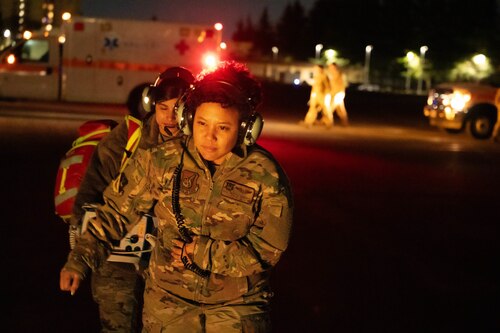 Two Airmen are walking towards the camera at night with medical equipment. An Ambulance is seen on the left side of of the photo.