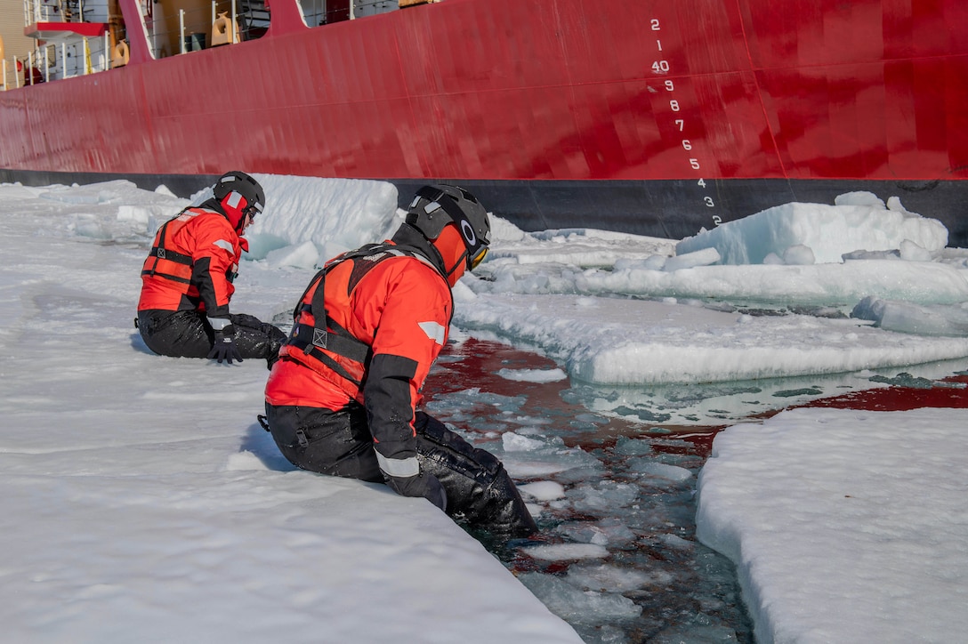 Two coast guardsmen wearing cold weather gear slide from a piece of broken ice into the semi-frozen water next to the side of a cutter.