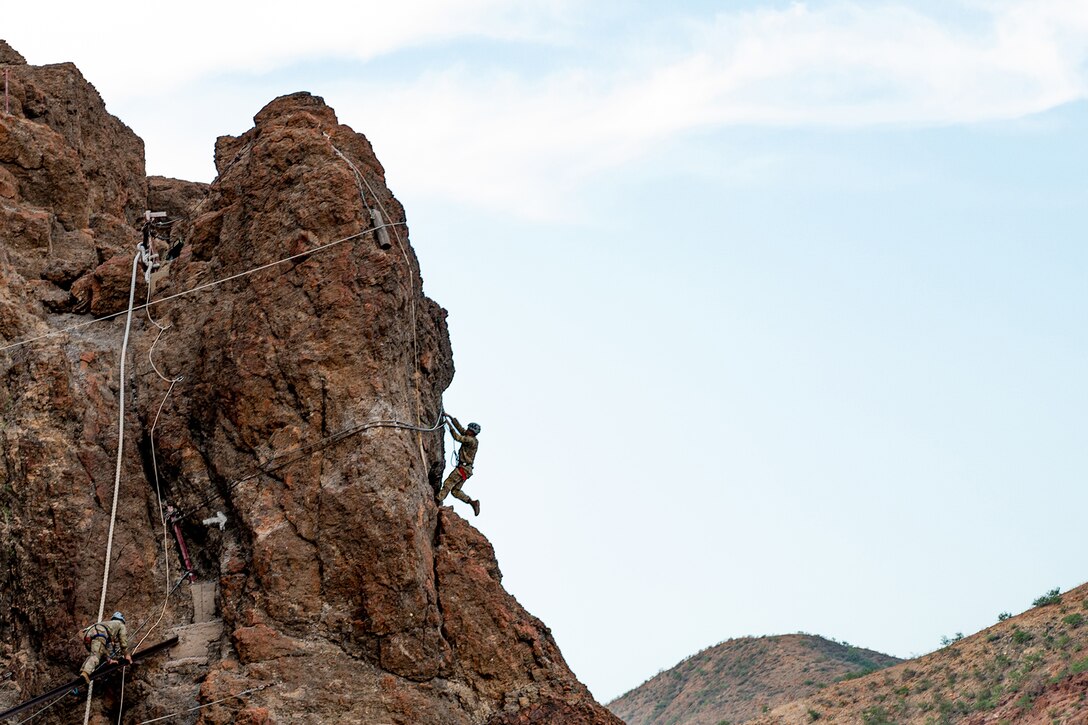 A soldier climbs a mountain during daylight as a second uses a climbing rope.