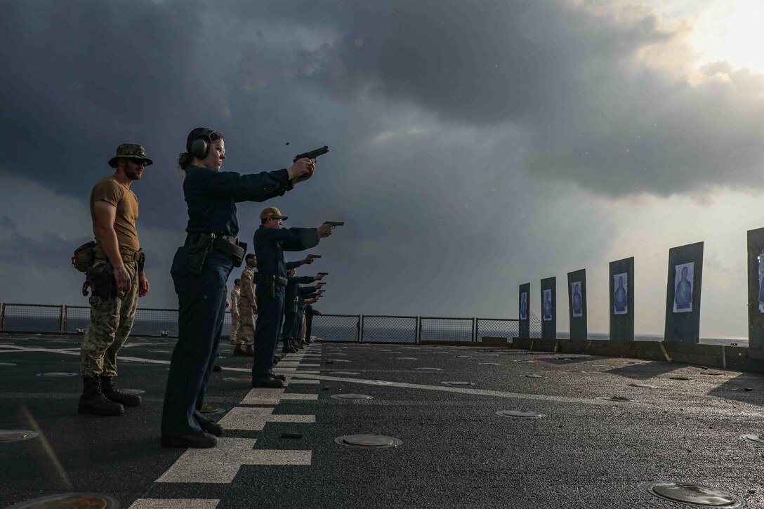 Sailors fire pistols at targets on the deck of a ship during a small arms qualification shoot on a cloudy day.