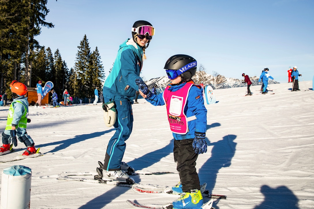 An instructor holds a military child’s hand while skiing as others ski down the snowy mountain in the background.