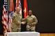 Male and female Soldier pose shaking hands in front of an array of flags