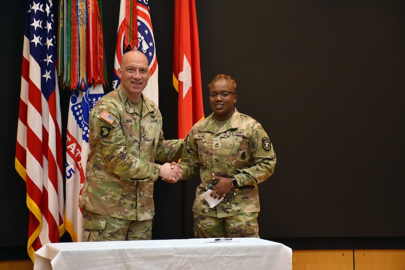 Male and female Soldier pose shaking hands in front of an array of flags