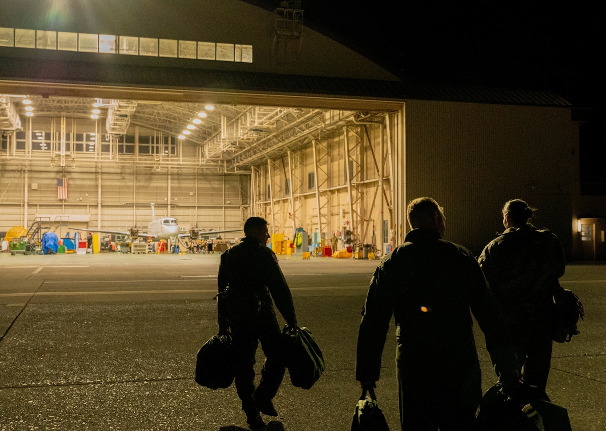 Three Airmen walk towards a hanger at night carrying their equipment. A C-12 aircraft is visible in the hanger.