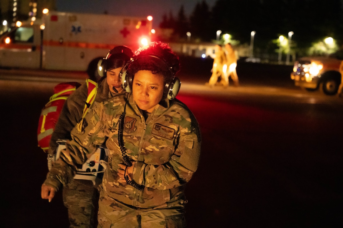 Two Airmen are walking towards the camera at night with medical equipment. An Ambulance is seen on the left side of of the photo.