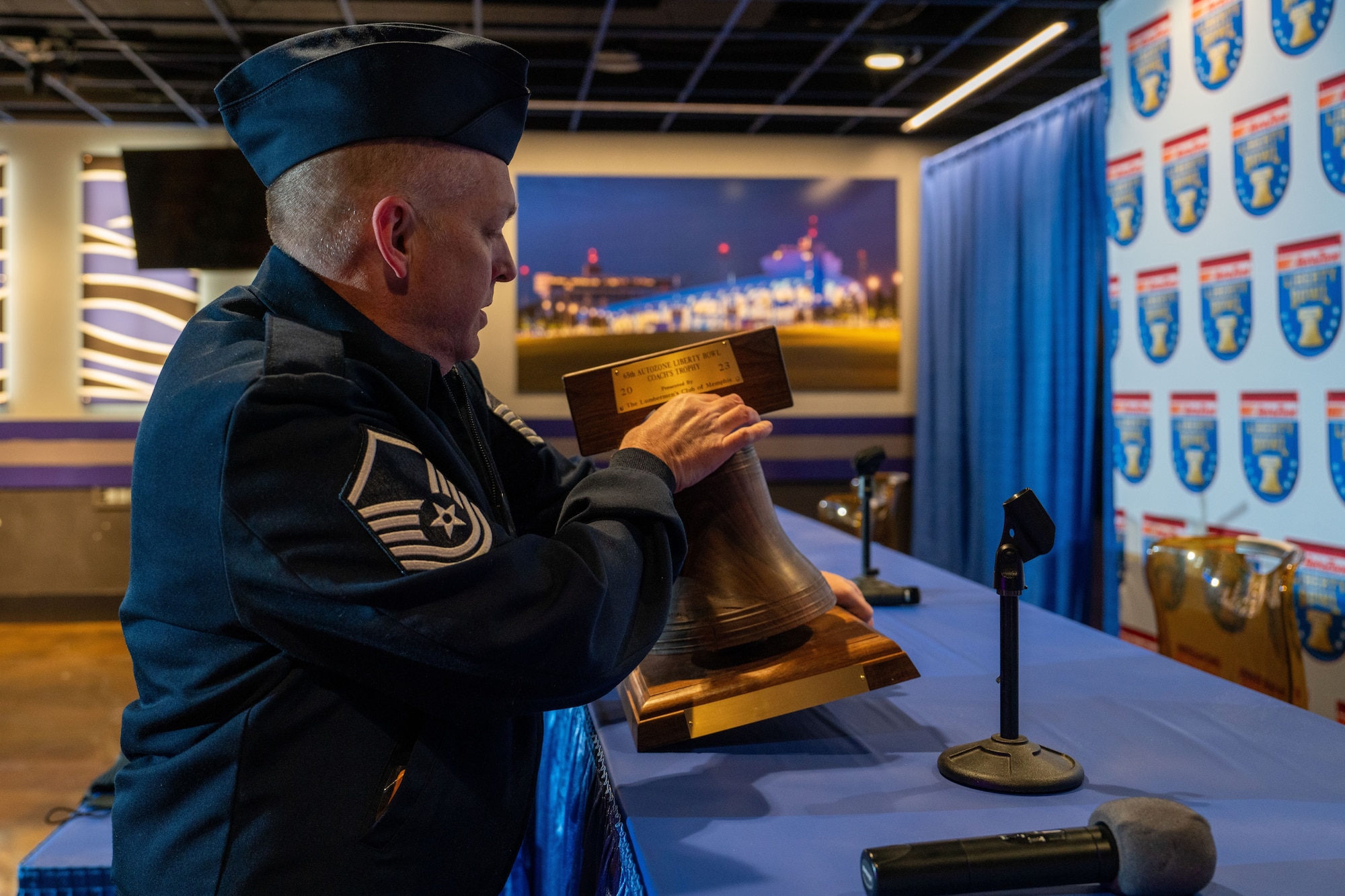 An image of a man in a U.S. Air Force dress uniform is inspecting or polishing a bell-shaped trophy on a presentation table, with the Liberty Bowl logos on a backdrop behind him.