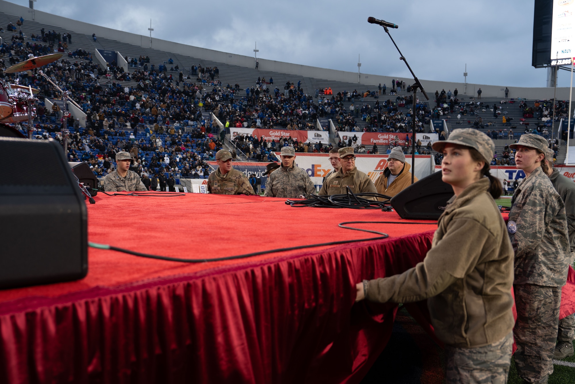 A photo of military personnel and civilians who are gathered around a red-covered stage on the football field, with various individuals looking in different directions.