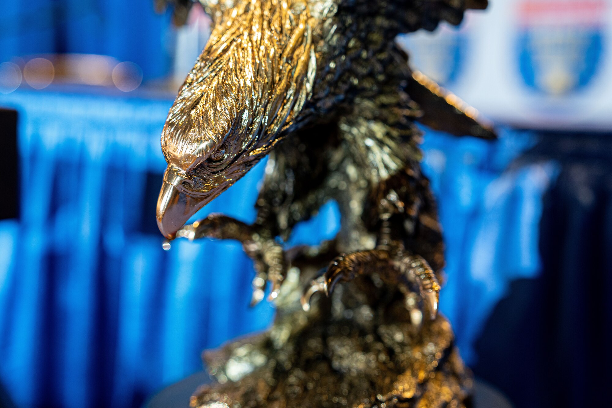 A close-up image of an eagle trophy. The trophy is in sharp focus against a blurred background featuring blue banners with the Liberty Bowl logo.
